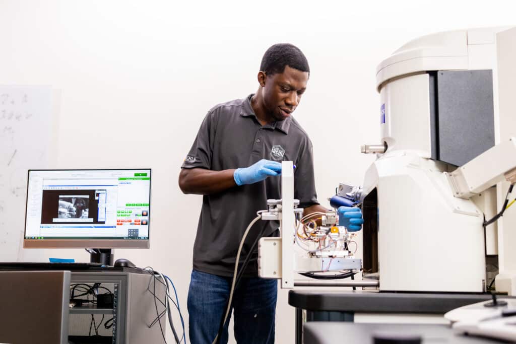 A black man works with a nano scale mechanical testing system integrated in a Scanning Electron Microscope inside an IAMM lab